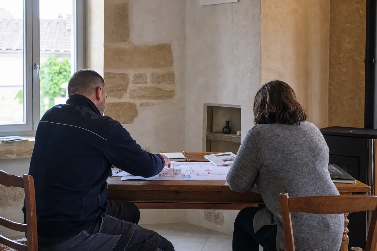 Silhouettes de dos d&rsquo;un artisan et d&rsquo;un client penchés sur un plan d&rsquo;installation de chauffage dans une maison en pierre de Bourgogne